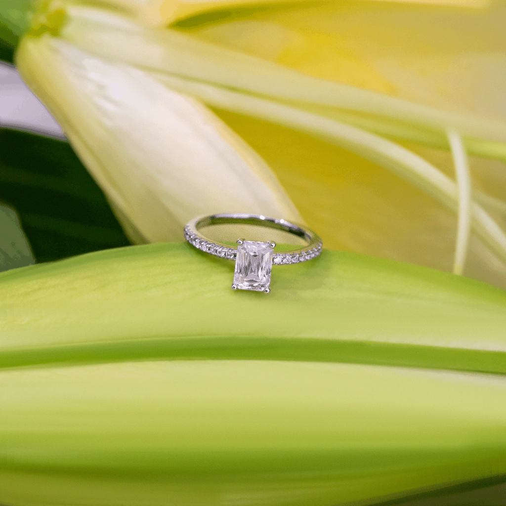A silver engagement ring with a lab-grown emerald-cut clear center stone and small side stones, resting on a bright green lily leaf with yellow petals in the background.